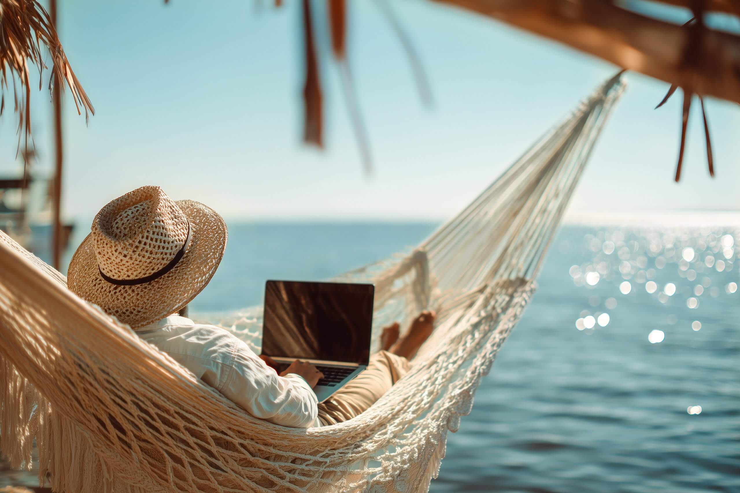 A man in a straw hat working on a laptop while relaxing in a white hammock on a tropical beach. Remote work, digital nomad lifestyle, and summer vacation concept by the blue ocean